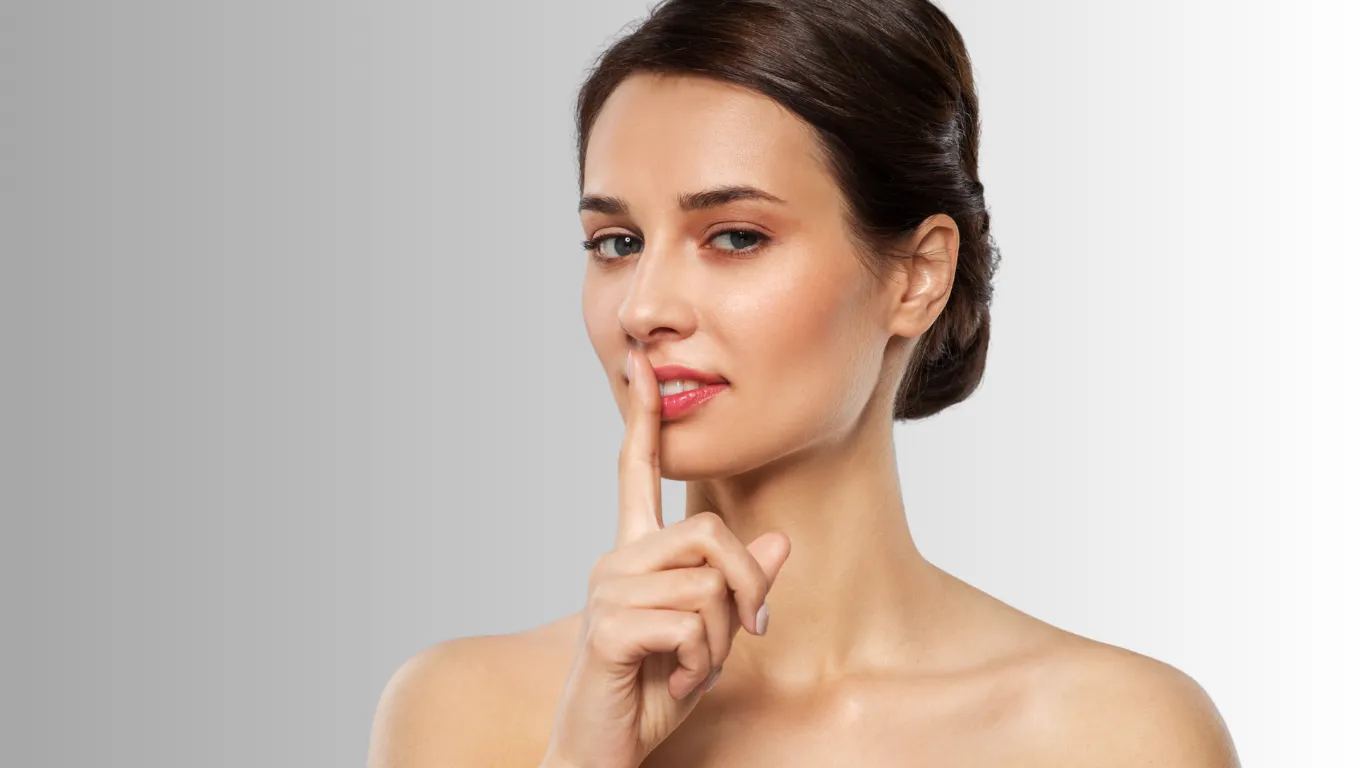 Woman with finger to her lips in a “shh” gesture, against a gray background